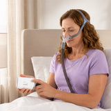 A woman in bed wears a nasal CPAP mask while adjusting pressure settings on the ResMed AirMini in her hand