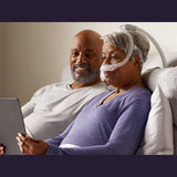 Two people sitting up in bed with a tablet, one of them wearing the DreamWear nasal pillow mask