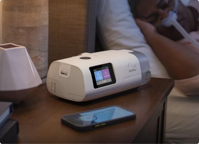 The ResMed AirCurve 11 VAuto on a bedside table, tubing running to a person asleep in a nasal mask