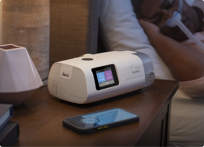 The ResMed AirCurve 11 VAuto on a bedside table, tubing running to a person asleep in a nasal mask